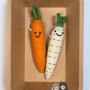 May include: Two carrot-shaped keychains with smiling faces and green tops rest in a brown paper container. One keychain is orange, the other is white with black stripes. The keychains are made of felt.
