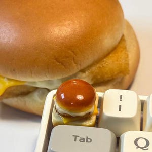 May include: A miniature burger with a glossy, golden-brown bun and a small patty, cheese, and bun. The burger is placed on a computer keyboard near the "Tab" key. A larger burger is in the background.