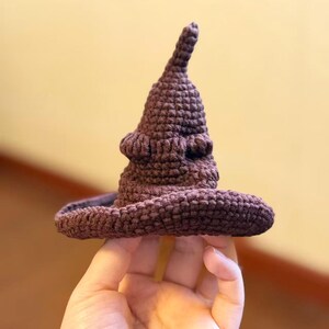 May include: A small, brown, crocheted hat with a pointed top and wide brim. The hat is held in a hand, showcasing its textured, handmade appearance. The hat's design is reminiscent of the Sorting Hat from the Harry Potter series.