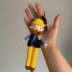 May include: A crocheted keychain figure depicting a person with a yellow hat, a brown beard, and a pipe. The figure wears a blue shirt, a dark navy vest with three small buttons, and yellow trousers with brown feet. A small yellow flower is attached to the vest.