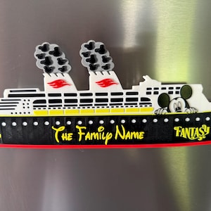 May include: A cruise ship magnet featuring a black hull with a yellow anchor, a white upper deck, and two smokestacks with grey smoke clouds. The text reads The Family Name and Fantasy, with a cartoon mouse character peering over the deck railing.