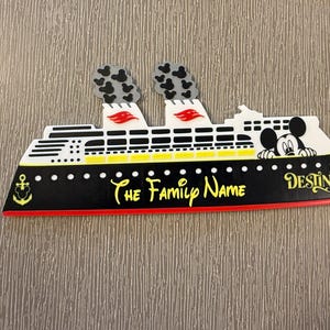 May include: A decorative cruise ship sign featuring a black hull with a red stripe, yellow accents, and two smokestacks with black Mickey Mouse head patterns. The text reads The Family Name and Destiny, with a Mickey Mouse character and anchor icon.