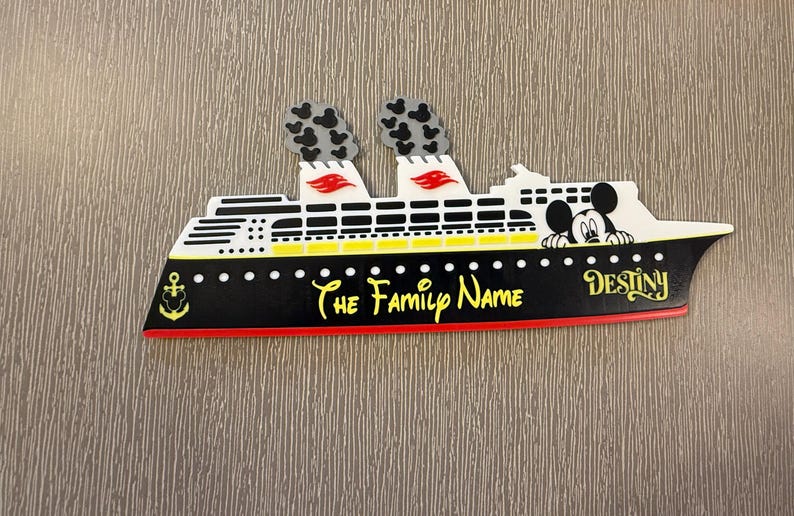 May include: A decorative cruise ship sign featuring a black hull with a red stripe, yellow accents, and two smokestacks with black Mickey Mouse head patterns. The text reads The Family Name and Destiny, with a Mickey Mouse character and anchor icon.