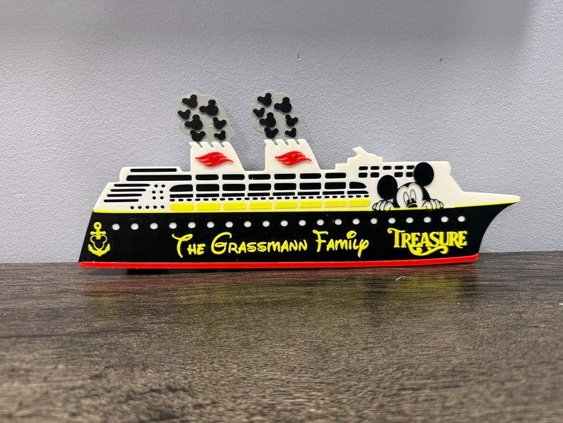 May include: A decorative cruise ship sign featuring a black hull with a red base, white upper decks, and two smokestacks with black Mickey Mouse head patterns. The text reads The Grassmann Family and Treasure, with a yellow anchor and Mickey icon.