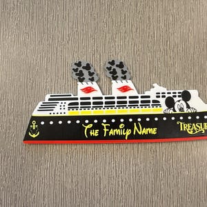 May include: A cruise ship magnet featuring a black hull with a red base, white upper decks, and two smokestacks with black Mickey Mouse head patterns. The text reads The Family Name and Treasure, with a Mickey Mouse character peeking over the deck.