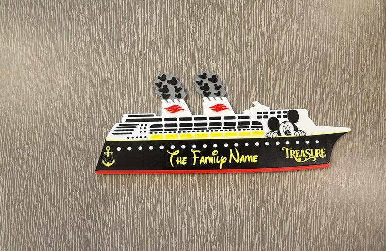 May include: A cruise ship magnet featuring a black hull with a red base, white upper decks, and two smokestacks with black Mickey Mouse head patterns. The text reads The Family Name and Treasure, with a Mickey Mouse character peeking over the deck.
