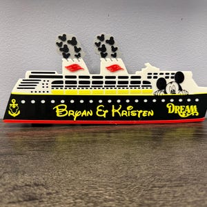 May include: A decorative cruise ship cutout featuring a black hull with a yellow anchor, the text Bryan & Kristen, and the word DREAM. The upper section is white with yellow accents, two smokestacks with black Mickey icons, and a Mickey Mouse head.