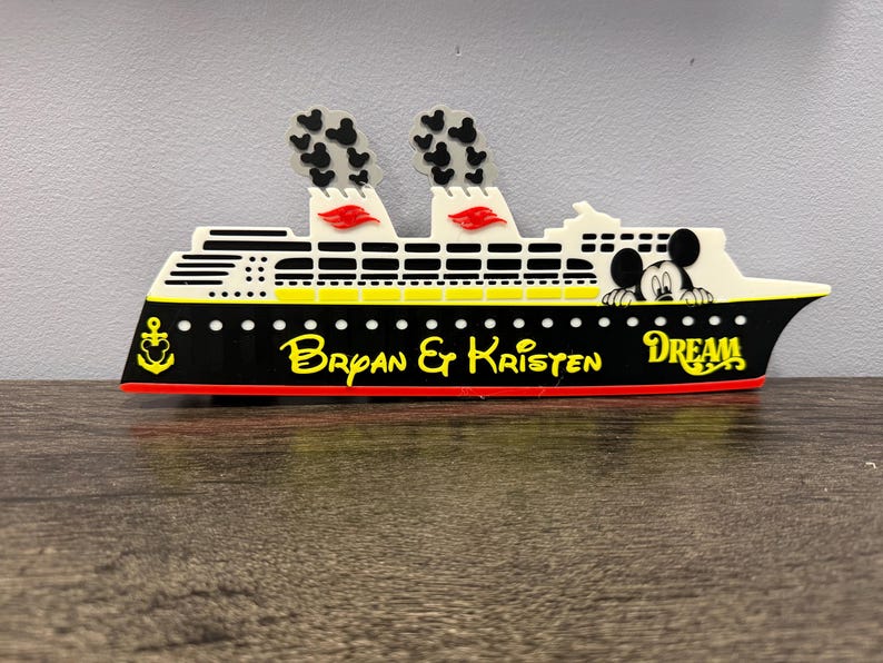 May include: A decorative cruise ship cutout featuring a black hull with a yellow anchor, the text Bryan & Kristen, and the word DREAM. The upper section is white with yellow accents, two smokestacks with black Mickey icons, and a Mickey Mouse head.