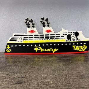 May include: A decorative cruise ship cutout featuring a black hull with a red stripe, white upper decks, and two smokestacks with black Mickey Mouse icon smoke. The ship displays the yellow text Perry and Fantasy, a yellow anchor icon, and a Mickey Mouse head.