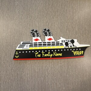 May include: A flat, cruise ship-shaped magnet featuring a black hull with a yellow stripe, white upper decks, and two smokestacks with grey Mickey Mouse icons. The text reads "The Family Name" and "WISH" in yellow, with a Mickey Mouse head graphic.