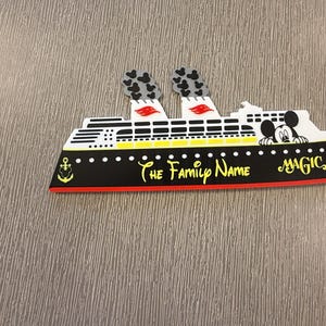 May include: A flat cruise ship decoration featuring a black hull with a yellow anchor icon, white superstructure, and two smokestacks with grey Mickey Mouse silhouettes. The text reads The Family Name and MAGIC, with a Mickey Mouse head peeking out.