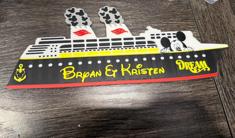 May include: A flat, decorative cruise ship magnet featuring a black hull with yellow text reading Bryan & Kristen and Dream. The white upper deck includes two smokestacks with Mickey Mouse head patterns and a graphic of Mickey Mouse peeking over.