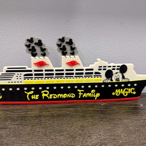 May include: A decorative cruise ship sign featuring a black hull with a yellow anchor, white upper decks, and two smokestacks emitting black Mickey Mouse-shaped smoke. The text reads The Redmond Family and Magic, with a Mickey Mouse character graphic.