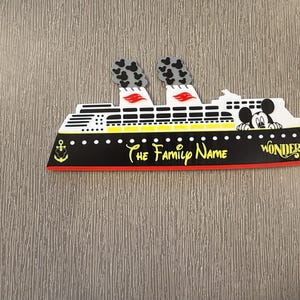 May include: A flat, cruise ship-shaped magnet featuring a black hull with a yellow anchor icon, white upper decks, and two smokestacks with black Mickey Mouse head patterns. The text reads The Family Name and WONDER in yellow script.