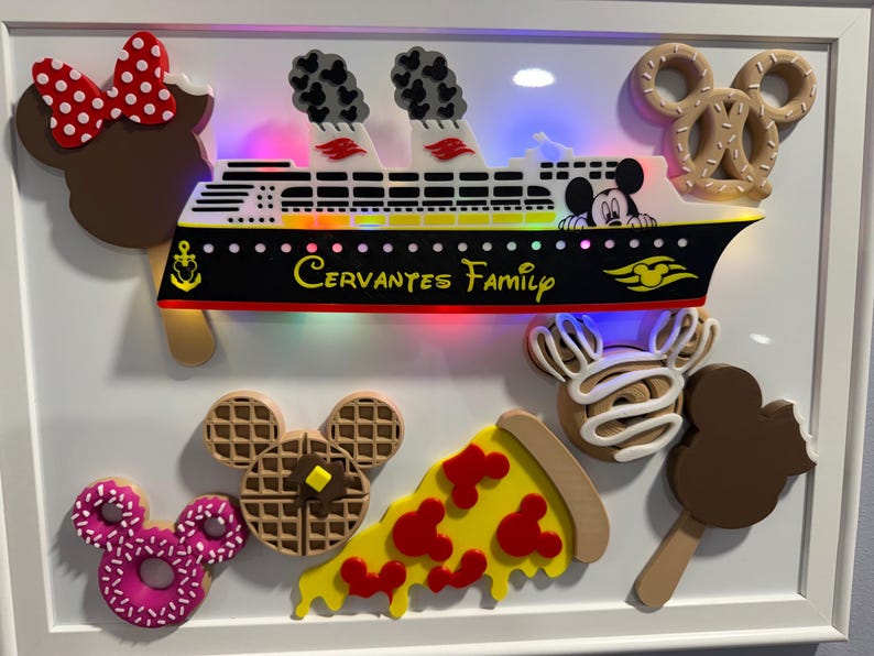 May include: A framed wall art piece featuring a light-up cruise ship model with the text "Cervantes Family" on the hull. Surrounding the ship are various food-themed icons including a chocolate bar, a pretzel, a waffle, pizza, and a donut.