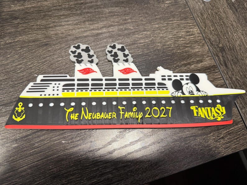 May include: A cruise ship magnet featuring a black hull with a yellow stripe, white upper decks, and two smokestacks with Mickey Mouse-shaped smoke. It displays the text "The Neubauer Family 2027" and "FANTASY" with a yellow anchor icon.