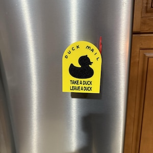 May include: A yellow fridge magnet with the words "DUCK MAIL" above a black duck silhouette. Below the duck is the text "TAKE A DUCK LEAVE A DUCK". The magnet is on a stainless steel fridge.