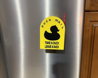 May include: A yellow fridge magnet with the words "DUCK MAIL" above a black duck silhouette. Below the duck is the text "TAKE A DUCK LEAVE A DUCK". The magnet is on a stainless steel fridge.