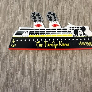 May include: A cruise ship magnet featuring a black hull with a yellow anchor icon, white upper decks, and two smokestacks with black Mickey Mouse head patterns. The text reads The Family Name and Adventure, with a Mickey Mouse head peeking out.