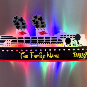 May include: A cruise ship magnet featuring a black hull with the text The Family Name and Fantasy in yellow. The white upper deck displays a Mickey Mouse character, two smokestacks with small Mickey icons, and red accents on the funnels.