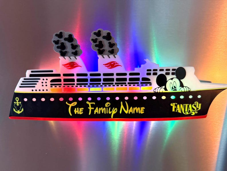 May include: A cruise ship magnet featuring a black hull with the text The Family Name and Fantasy in yellow. The white upper deck displays a Mickey Mouse character, two smokestacks with small Mickey icons, and red accents on the funnels.