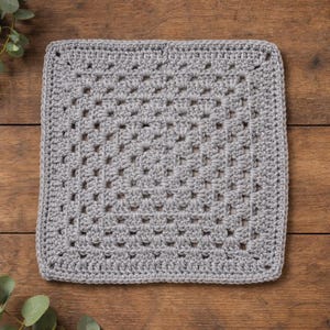 Op de afbeelding: Een vierkant, lichtgrijs gehaakt item. Het heeft een traditioneel granny square-ontwerp met een gestructureerd, open weefpatroon. De randen zijn netjes afgewerkt. Het item wordt gepresenteerd op een houten oppervlak.