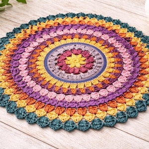 Op de afbeelding: Een kleurrijke, ronde gehaakte doily met een bloemenontwerp. De doily heeft concentrische cirkels in verschillende kleuren, waaronder teal, geel, oranje, roze en paars. Het midden heeft een bloemachtig patroon. De doily ligt op een licht houten oppervlak. Diameter ca. 30 cm.