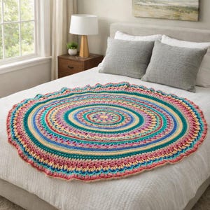 以下が含まれることがあります： ターコイズ、ピンク、イエロー、ブルーの色合いで同心円を描いた、カラフルな丸いかぎ針編みのラグ。 縁がスカラップになったラグは白いベッドの上に置かれています。 デザインはマンダラを連想させ、直径は約120cmです。