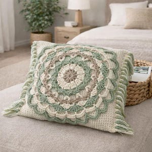 Puede incluir: Un cojín cuadrado de ganchillo color crema con un diseño floral circular en tonos verdes y beige. El cojín tiene un borde con flecos y está colocado sobre una superficie de tono neutro, ideal para la decoración del hogar.
