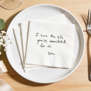 Könnte beinhalten: Ein weißer Teller mit einem Stapel weißer Servietten, einer Gabel und einem Messer. Die obere Serviette trägt den handgeschriebenen Text: "Ich liebe das Leben, das du für uns manifestiert hast. Eric."
