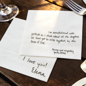Op de afbeelding: Twee witte papieren servetten met handgeschreven tekst in zwarte inkt. Eén servet leest "I'm overwhelmed with gratitude...Grace of God." Een ander zegt "I love you! Elena." Een vork en mes zijn zichtbaar.