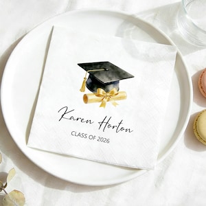 Può includere: Tovagliolo quadrato bianco con un'illustrazione di cappello di laurea e diploma. Il testo "Karen Horton" e "CLASS OF 2026" è stampato in nero. Il tovagliolo è su un piatto bianco, con due macarons e un bicchiere d'acqua.