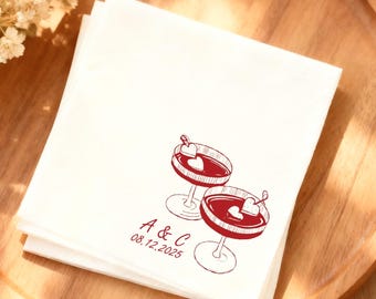 Peut inclure: Une pile de serviettes à cocktail blanches avec une illustration rouge de deux verres à martini. Chaque verre a une garniture en forme de cœur. Le texte "A & C 08.12.2025" est imprimé en dessous. Les serviettes sont sur un plateau en bois.