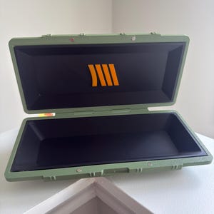 以下が含まれることがあります： 黒い内装の、オリーブグリーンの長方形のケース。ケースにはヒンジ付きの蓋と、内側に明るいオレンジ色のロゴが付いています。ケースは白い表面に置かれています。