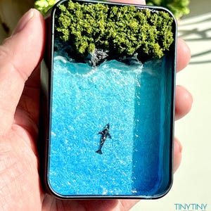 May include: Miniature ocean diorama in a small, rectangular tin. The scene features a turquoise ocean with a small black whale, a rocky shore, and green foliage. The diorama is held in a hand. The text "TINY TINY WORLD" is visible.