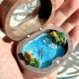 May include: A small oval wooden box with a hinged lid containing a miniature diorama of a blue ocean scene with a whale, green foliage, and rocky elements. The text TINY TINY WORLD is printed in blue on the lower right side of the image.