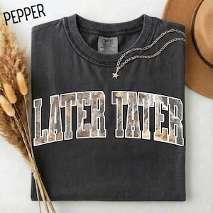 May include: A dark gray t-shirt with the text "LATER TATER" in a camouflage pattern. The shirt has a crew neck and short sleeves. A silver necklace with a star pendant is on the shirt. The word "PEPPER" is in the upper left corner.