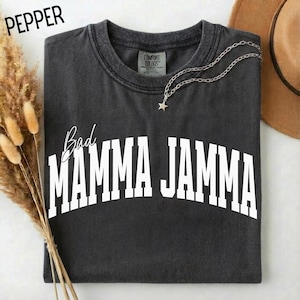 May include: A dark gray t-shirt with the words "Bad Mamma Jamma" in white block letters. The shirt has a crew neck and a small silver necklace with a star pendant. The word "Pepper" is in the upper left corner.