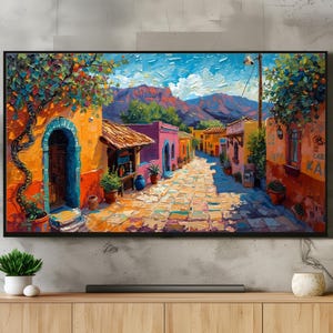 May include: A framed oil painting depicts a vibrant street scene with colorful buildings and a cobblestone path. The artwork features a warm color palette with orange, pink, and blue hues, evoking a sense of a sunny day. The painting is displayed above a wooden cabinet.
