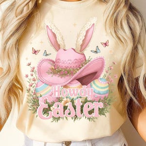 May include: A light yellow t-shirt featuring a pink cowboy hat with bunny ears, decorated with flowers and butterflies. The text "Howdy Easter" is displayed in pink script below the hat and Easter eggs.