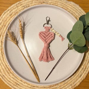 Peut inclure: Un porte-clés en macramé rose avec un motif en forme de cœur et une gland. Le porte-clés porte le mot "MAMAN" épelé en perles de bois. Le porte-clés est sur une assiette blanche avec des épis de blé et de la verdure.