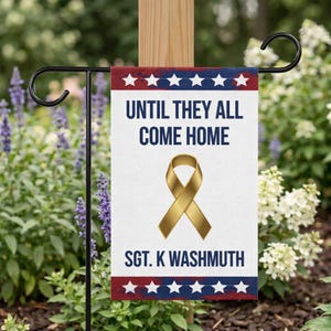 May include: A garden flag with a white background, red and blue borders, and the text "UNTIL THEY ALL COME HOME." A gold ribbon graphic is centered below the text. The name "SGT. K WASHMUTH" is printed at the bottom.