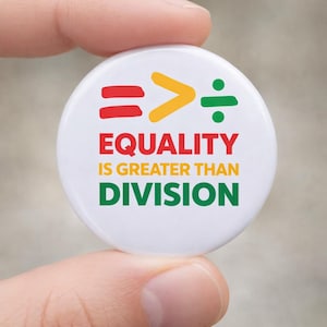 May include: A white button with the text "EQUALITY IS GREATER THAN DIVISION" in red, yellow, and green. The button also features mathematical symbols: an equals sign, a greater than sign, and a division symbol.