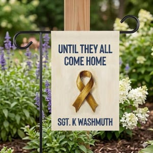 May include: A beige garden flag with navy blue text reading "UNTIL THEY ALL COME HOME". A gold ribbon graphic is below the text, with "SGT. K WASHMUTH" at the bottom. The flag hangs on a black metal stand, surrounded by greenery.