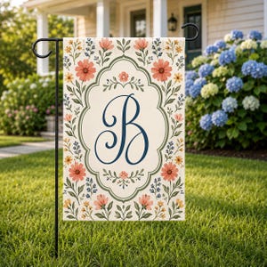 May include: A garden flag featuring a floral design with a large, navy blue letter "B" in the center. The flag has a cream-colored background and is decorated with peach and yellow flowers, and green leaves. The flag is displayed on a black metal stand.