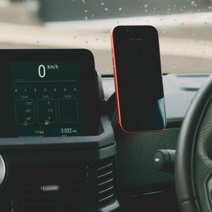 Puede incluir: Un salpicadero de coche con una pantalla digital que muestra 0 km/h y otra información del vehículo. Un smartphone con marco rojo está montado en el salpicadero. La fecha en el teléfono es "Mon 25 May".