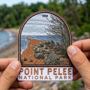 May include: Embroidered patch featuring a coastal landscape with the text "POINT PELEE NATIONAL PARK" and the year "1918". The patch depicts a beach, rocks, and trees, with a blue and white sky. The patch is held by a person.