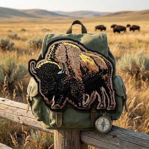May include: An olive green backpack with a large embroidered bison patch. The patch features brown, black, and yellow details. A vintage compass hangs from the wooden fence in the foreground, with a field and bison in the background.