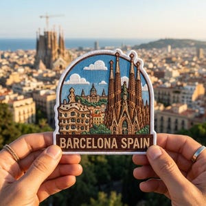 May include: An embroidered patch featuring a detailed depiction of the Sagrada Familia and other Barcelona architecture against a light blue sky with white clouds. The patch has a brown border and the text BARCELONA SPAIN in brown capital letters.