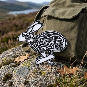 May include: A white and black embroidered patch featuring a rabbit design with Celtic knot patterns. The patch is placed on a rock with a green backpack in the background. The rabbit design has intricate details and a stylized appearance.
